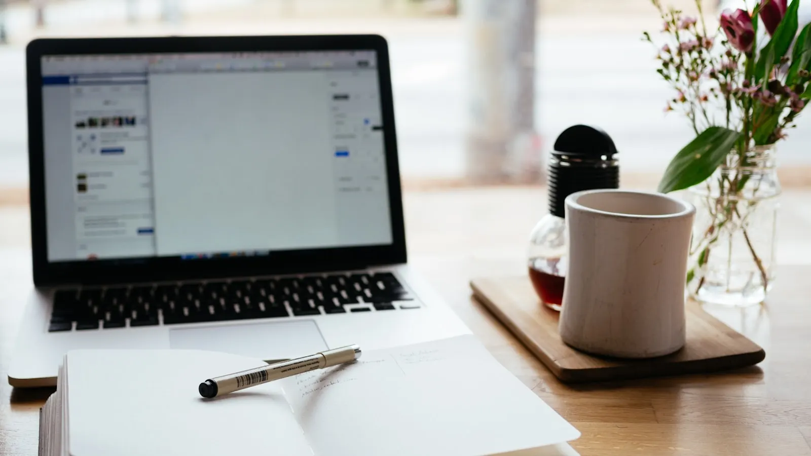 Ein Laptop und ein offenes Notizbuch mit einem Stift auf einem Holzschreibtisch, die die Arbeit des Schreibens und Verfeinerns eines KI-Telefonassistenten-Prompts darstellen