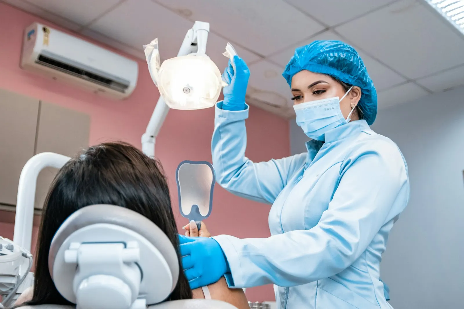 Un professionista dentale mascherato in camice che regola la luce sopraelevata e tiene uno specchietto dentale accanto a un paziente sdraiato sulla poltrona
