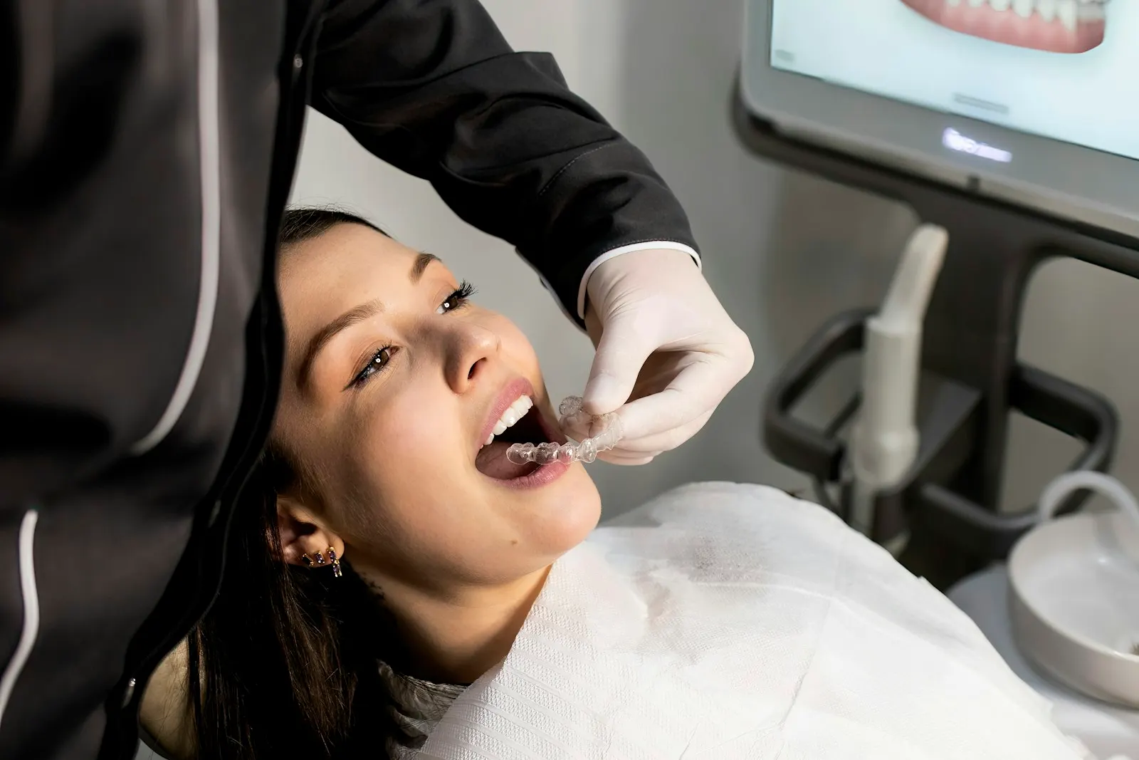 Ein Zahnarzt passt einem Patienten, der im Behandlungsstuhl liegt, eine durchsichtige Zahnschiene an, während auf dem Bildschirm dahinter ein intraoraler Scan der Zähne zu sehen ist.
