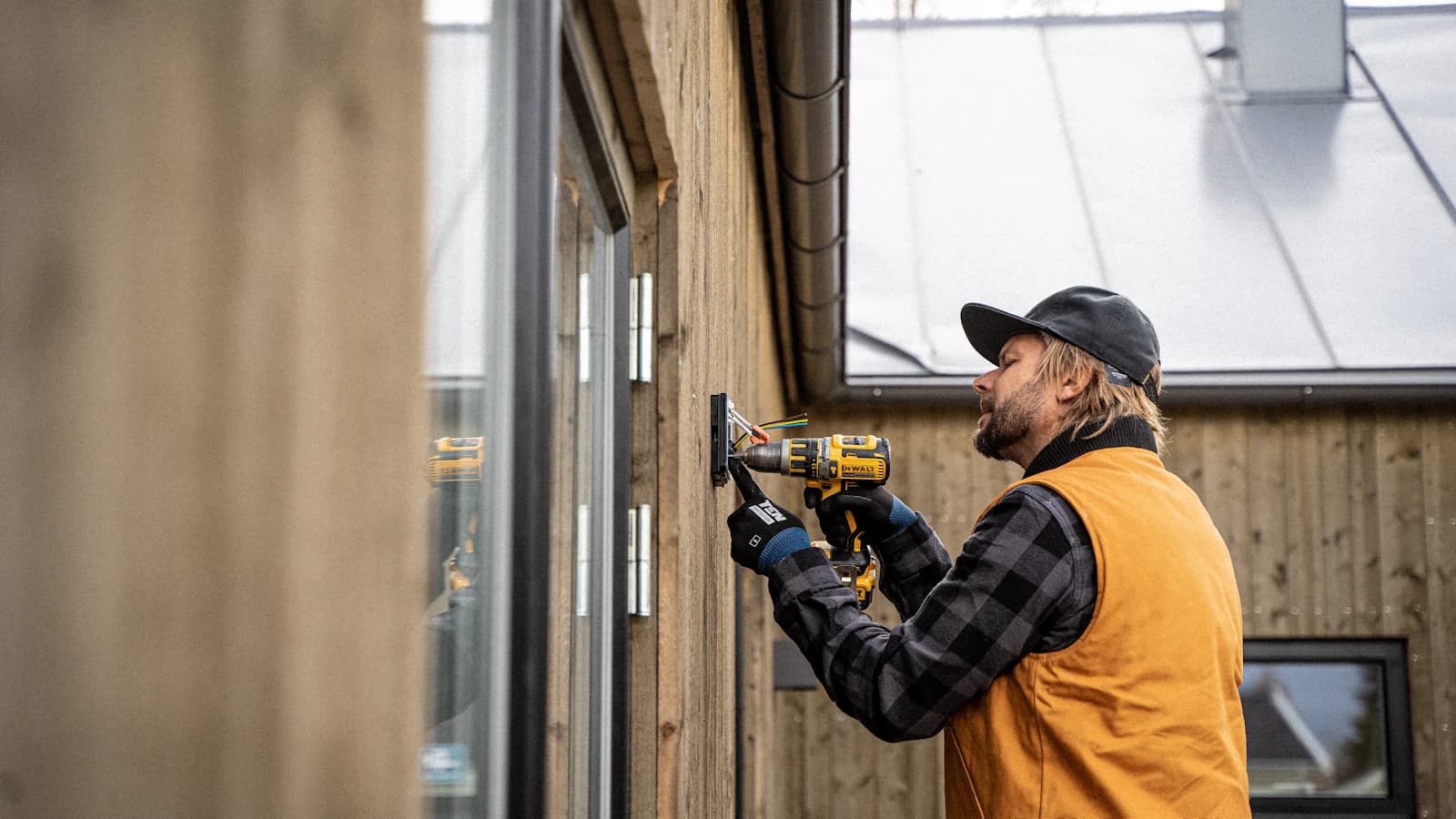 Ein Elektriker mit schwarzer Kappe und orangefarbener Arbeitsweste installiert mit einem Akkuschrauber eine Außensteckdose an einem Holzhaus, beide Hände bei der Arbeit, während das Telefon woanders klingelt