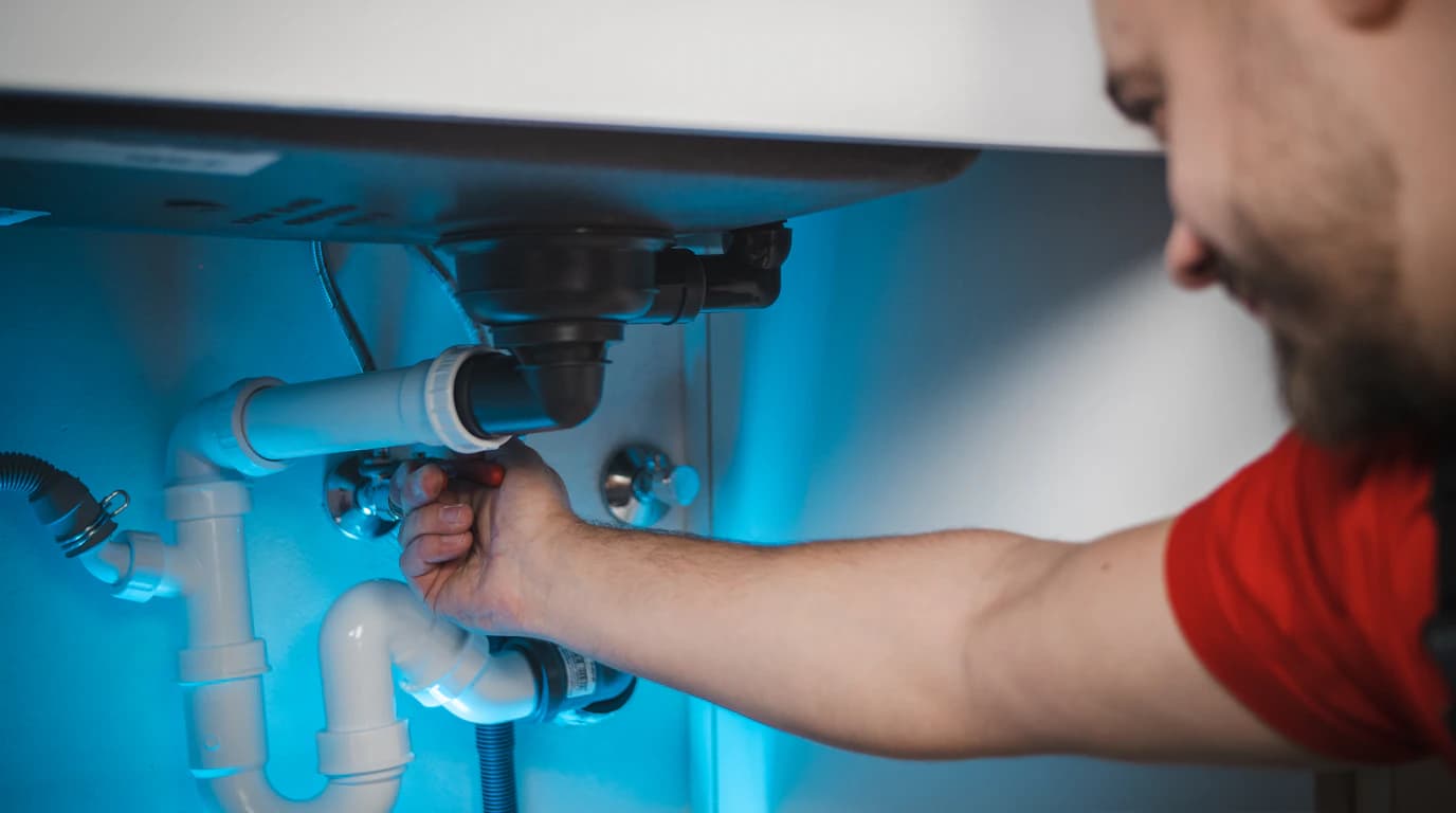 Ein Klempner greift unter eine Spüle, um den Siphon und die Zuleitungen zu reparieren, beide Hände sind im Schrank beschäftigt