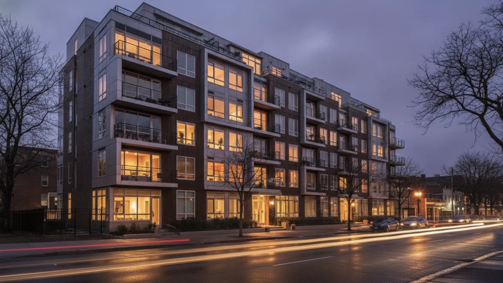 A mid-rise apartment building at dusk with warm lights in the windows - the tenant calls a property management answering service covers after hours